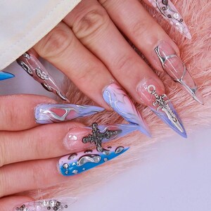 May include: Close-up of long, pointed, blue and pink nail art with silver and black accents. The nails feature a cross, a sword, and swirls.
