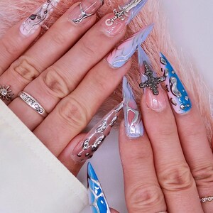 May include: Close-up of long, pointed, blue and white painted fingernails with silver accents. Some nails have small silver charms attached. The nails are on a hand wearing a silver ring with the word "always" on it.