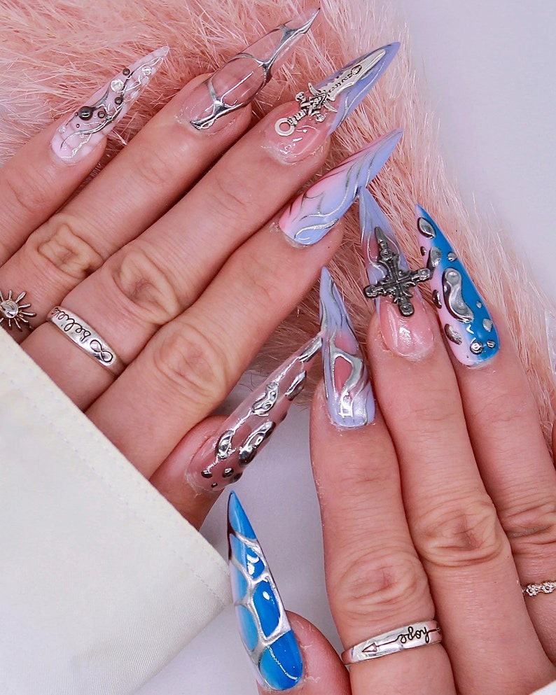 May include: Close-up of long, pointed, blue and white painted fingernails with silver accents. Some nails have small silver charms attached. The nails are on a hand wearing a silver ring with the word "always" on it.