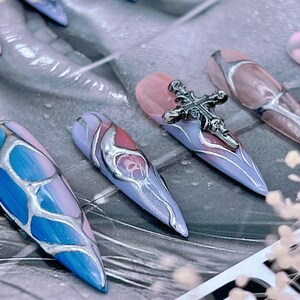 May include: Close-up of several long, pointed, false nails with a silver cross charm on one nail. The nails are painted in a blue and pink marble design with silver accents.