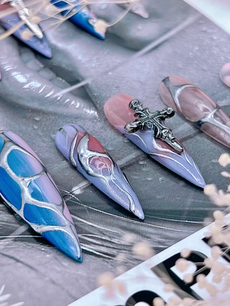 May include: Close-up of several long, pointed, false nails with a silver cross charm on one nail. The nails are painted in a blue and pink marble design with silver accents.