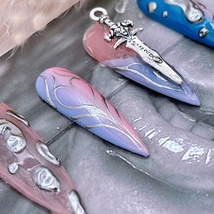 May include: A set of long, pointed, false nails with a gradient design. The nails are painted in shades of pink and blue with silver accents. One nail has a silver sword charm attached to it.