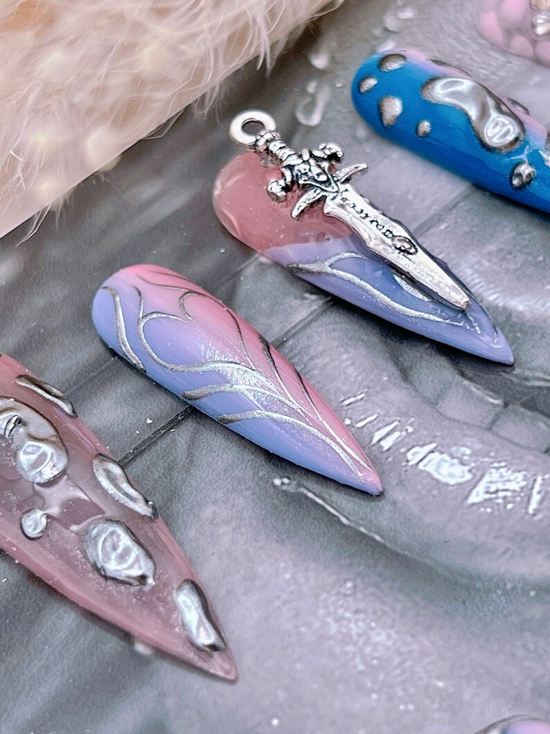 May include: A set of long, pointed, false nails with a gradient design. The nails are painted in shades of pink and blue with silver accents. One nail has a silver sword charm attached to it.