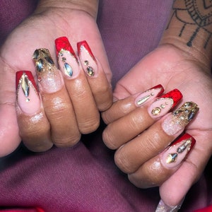 May include: Close-up of two hands with long, red and gold decorated nails. The nails are a long, almond shape and have a red base with gold glitter and rhinestones.