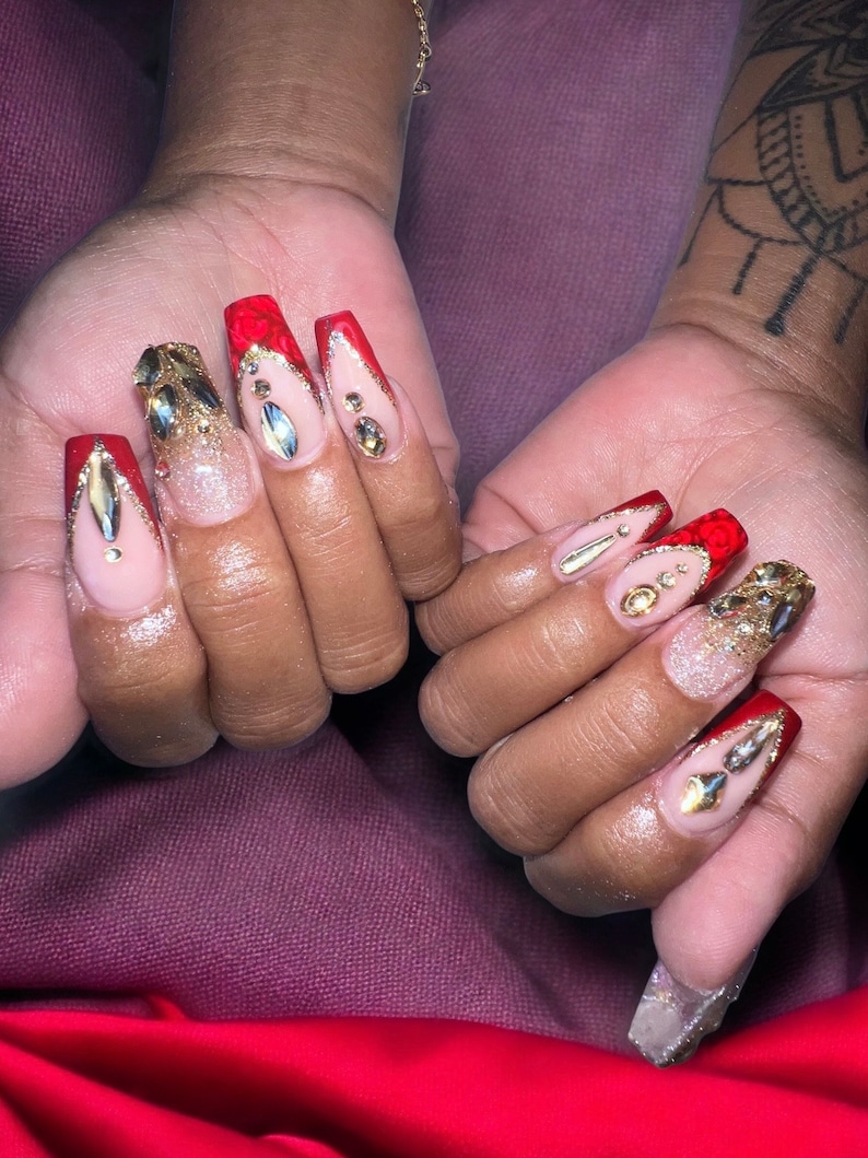 May include: Close-up of two hands with long, red and gold decorated nails. The nails are a long, almond shape and have a red base with gold glitter and rhinestones.