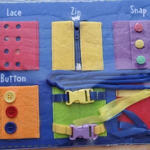 Peut inclure: Un tableau en feutre bleu avec différentes textures de tissu et activités pour enfants. Le tableau présente un carré rouge avec des trous pour lacer, un carré jaune avec une fermeture éclair, un carré violet avec des boutons-pression, un carré orange avec des boutons et un carré vert avec une boucle.