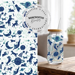 Könnte beinhalten: Ein durchsichtiger Glasbecher mit Bambusdeckel und Glasstrohhalm. Der Becher hat ein blaues und türkisfarbenes Weltraumthema mit Hunden, Planeten und Sternen. Das Design wiederholt sich im Hintergrund.