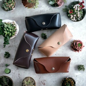 May include: Four different leather eyeglass cases in various colours, including brown, black, and beige. Each case has a snap closure and a curved shape.