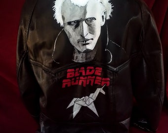May include: Black leather jacket with a hood, featuring a hand-painted portrait and the word "WANTED" on the back. Below the portrait, the jacket displays the text "BLADE RUNNER" and a white origami unicorn.