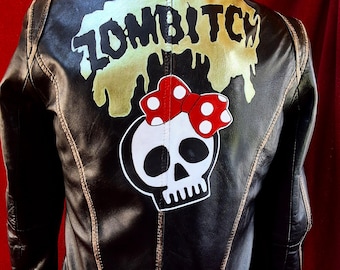 Könnte beinhalten: Schwarze Lederjacke mit einem goldenen "ZOMBIBITCH"-Design und einem Totenkopf mit einer roten Schleife mit weißen Punkten.