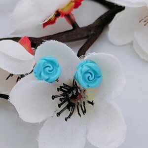 Può includere: Un paio di orecchini a forma di rosa blu. Gli orecchini sono su uno sfondo bianco con fiori bianchi.