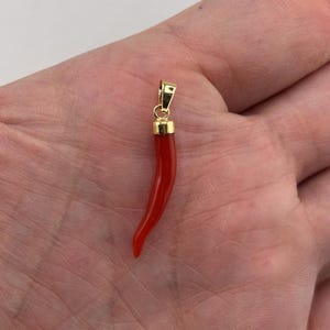 May include: A red chilli pepper pendant with a gold-coloured bail. The pendant is a vibrant red colour and has a smooth, curved shape. The bail is a rectangular shape with a small loop for a chain.