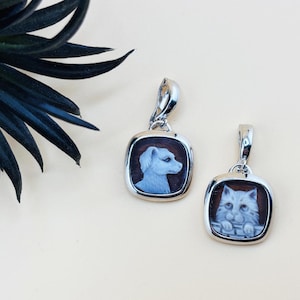 Könnte beinhalten: Zwei silberne Anhänger mit quadratischen Rahmen, jeweils mit einem Cameo eines Haustiers. Einer zeigt ein Hundeportrait, der andere eine Katze. Die Cameos sind hellblau auf braunem Hintergrund.