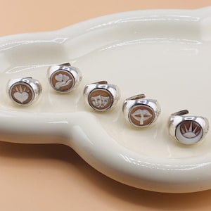 Puede incluir: Un conjunto de anillos de plata con diversos diseños en relieve. Cada anillo presenta un símbolo diferente, incluyendo un corazón, manos sosteniendo corazones, máscaras de teatro, labios y un sol y una luna. Los anillos se exhiben en un plato blanco de forma ovalada.