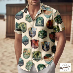 May include: A cream-coloured short-sleeved shirt with multiple illustrated badges. The badges feature text like "Aragorn" and "The Shire" in green, blue, and red. The shirt has a button-down collar and is worn by a person.
