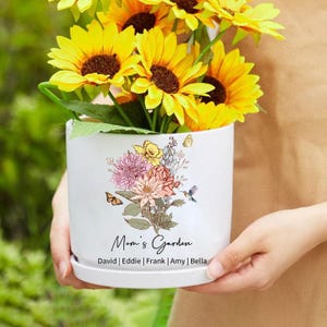 May include: A white ceramic flower pot with a floral design and the text "Mom's Garden." The pot holds bright yellow sunflowers. The pot also has the names David, Eddie, Frank, Amy, and Bella.