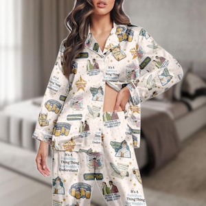 Può includere: Pigiama color crema con un motivo ripetuto di illustrazioni e testo relativi al musical "Hamilton". La maglia a maniche lunghe e i pantaloni presentano immagini di personaggi, citazioni ed elementi scenici. Il testo include frasi come "Showtime" e "It's a Hamilton Thing."