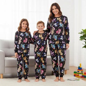 May include: Three people wearing matching black pajama sets adorned with colourful cartoon pony characters. Each set comprises a long-sleeved top and trousers. The image suggests a family or group. The background includes a sofa, rug, and children's toys.