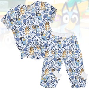 Può includere: Un completo pigiama per bambini composto da due pezzi. Presenta un motivo ripetuto di cani dei cartoni animati, fiori blu e foglie su sfondo bianco. Il set include una maglietta a maniche corte e pantaloni abbinati con polsini e vita bianchi.