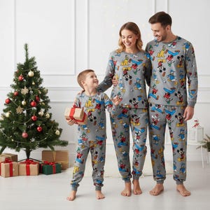 May include: Matching gray pajamas featuring Mickey Mouse and friends. The long-sleeved tops and pants are worn by a family of three. A Christmas tree and wrapped gifts are in the background, suggesting a holiday theme.
