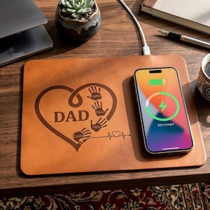 May include: A rectangular tan leather wireless charging pad featuring a laser-engraved design of a heart, the word DAD, four handprints with the names Sophie, Ethan, Mia, and Liam, and a heartbeat line. A smartphone is charging on the pad.