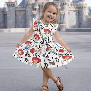 Puede incluir: Un vestido blanco con un estampado repetido de un personaje de vaquera de dibujos animados con sombrero rojo y atuendo azul, y manchas negras. El vestido tiene mangas cortas con volantes. La niña lleva sandalias marrones.