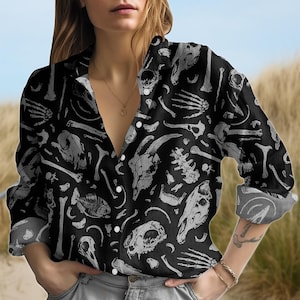 Könnte beinhalten: Schwarzes Button-Down-Hemd mit einem sich wiederholenden Muster aus grauen Tierschädeln, Knochen und Skelettelementen. Das langärmlige Hemd hat einen klassischen Kragen und einen Knopfverschluss. Das Hemd ist aus leichtem Stoff gefertigt.