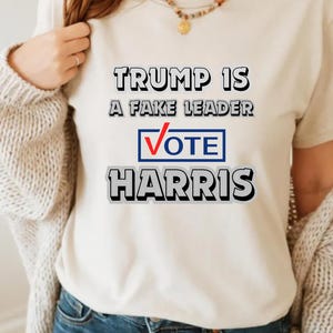 May include: A white t-shirt with the text "Trump is a fake leader Vote Harris" printed on it in black, red, and blue.