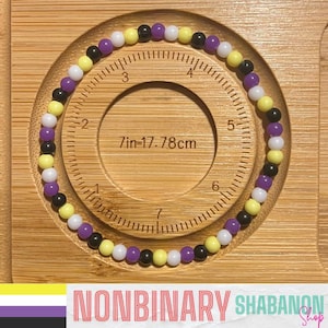 Könnte beinhalten: Ein Nonbinary-Pride-Armband mit Perlenmuster. Das Armband hat ein Muster aus weißen, gelben, lila und schwarzen Perlen. Das Armband wird auf einer Holzoberfläche mit einem kreisförmigen Ausschnitt und einem Lineal in cm angezeigt.
