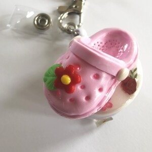 Könnte beinhalten: Ein pinkfarbener Miniatur-Clog-Schlüsselanhänger mit roter Blume und grünem Blattakzent. Der Schlüsselanhänger enthält einen Metallverschluss, einen Schlüsselring und einen transparenten Kunststoffausweishalter. Der Clog ist mit einer Blume und einer Rose verziert.