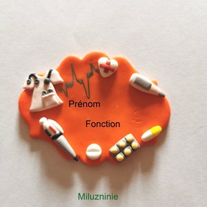 May include: Orange clay name tag with a stethoscope, a heart with a cross, a thermometer, a pen, pills, and a pill bottle. The tag says "Prénom" and "Fonction".