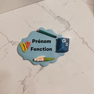 May include: A light blue cloud-shaped plaque featuring the black text Prénom Fonction. The plaque is decorated with a green pencil, a blue book with the letters a and z, a yellow paper clip, and a white and green pen attached to the surface.