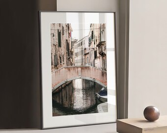 May include: Framed print of a canal scene in Venice, Italy. The image features a stone bridge over a canal with buildings on either side. A small boat is docked in the water. The print is in a black frame.