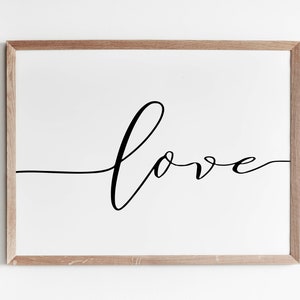 Puede incluir: Una impresión enmarcada con la palabra "amor" en elegante escritura negra. La palabra está centrada en un fondo blanco, y el marco es de madera marrón claro. Un diseño minimalista.