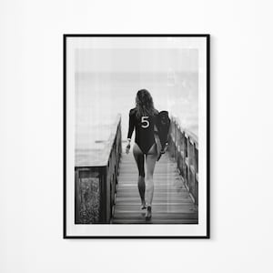 May include: A black and white photograph of a person walking on a wooden pier towards the sea, carrying a surfboard with the number "5" on it.
