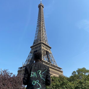 Puede incluir: Una persona con una chaqueta negra con las palabras "DARE OR DIE" pintadas en la espalda, de pie frente a la Torre Eiffel en París, Francia.