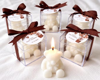 May include: Five bear-shaped candles, one lit and four inside clear square boxes. Each box is tied with a brown ribbon and a circular tag reading JULIA'S BABY SHOWER 21.12.2027 with a hot air balloon illustration. The candles are off-white.
