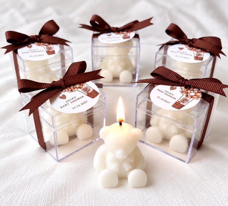 May include: Five bear-shaped candles, one lit and four inside clear square boxes. Each box is tied with a brown ribbon and a circular tag reading JULIA'S BABY SHOWER 21.12.2027 with a hot air balloon illustration. The candles are off-white.