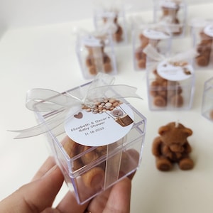以下が含まれることがあります： 透明なプラスチック製の箱の中に茶色のぬいぐるみが入っています。箱は銀色のリボンで結ばれており、丸いラベルには「Elizabeth & Oscar's Baby Shower 11.18.2023」と書いてあります。