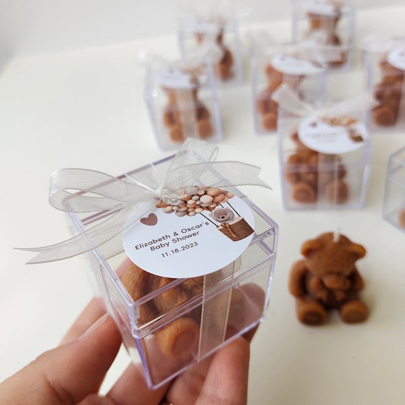 以下が含まれることがあります： 透明なプラスチック製の箱の中に茶色のぬいぐるみが入っています。箱は銀色のリボンで結ばれており、丸いラベルには「Elizabeth & Oscar's Baby Shower 11.18.2023」と書いてあります。