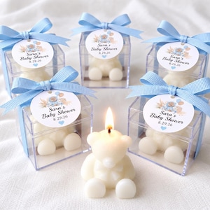 May include: Five clear square boxes containing white bear-shaped candles, each topped with a light blue ribbon and a circular tag reading Sara's Baby Shower 8.29.26. One lit white bear-shaped candle sits in the front center without a box.