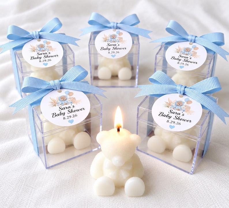 May include: Five clear square boxes containing white bear-shaped candles, each topped with a light blue ribbon and a circular tag reading Sara's Baby Shower 8.29.26. One lit white bear-shaped candle sits in the front center without a box.