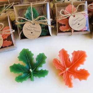 Puede incluir: Velas en forma de hoja en verde y naranja, algunas empaquetadas en cajas transparentes atadas con hilo. Cada caja tiene una etiqueta que dice "Simran's Bridal Shower" y la fecha 12.21.2021. Las velas están sobre una superficie blanca.