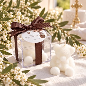 May include: A clear square plastic box containing white bear-shaped candles, tied with a brown ribbon and a circular tag reading JULIA BABY SHOWER 21.12.2023. A single white bear-shaped candle sits outside the box next to the gift packaging.
