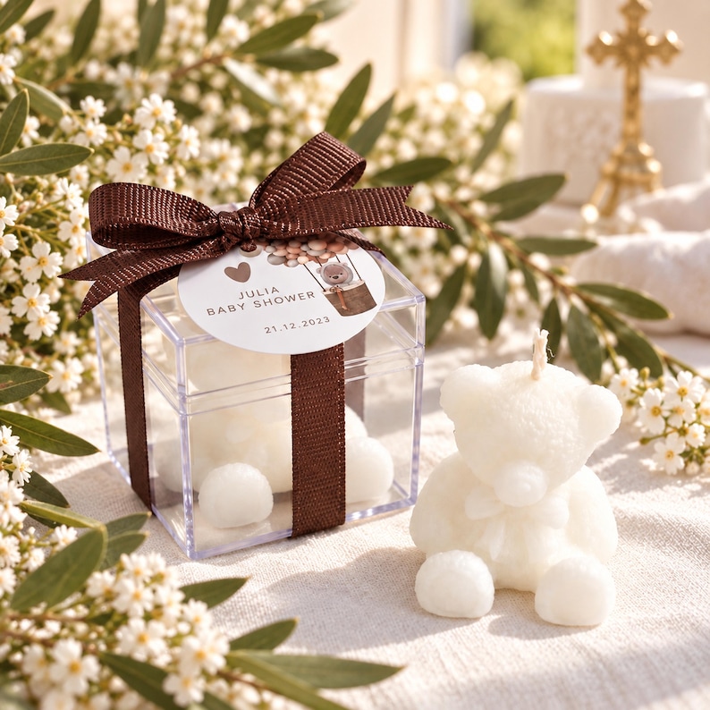May include: A clear square plastic box containing white bear-shaped candles, tied with a brown ribbon and a circular tag reading JULIA BABY SHOWER 21.12.2023. A single white bear-shaped candle sits outside the box next to the gift packaging.
