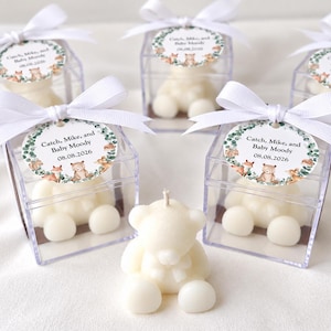 May include: Five off-white bear-shaped candles, four of which are enclosed in clear square plastic boxes topped with white ribbon bows and circular tags featuring woodland animal illustrations and the text Catch, Mike, and Baby Moody 08.08.2026.