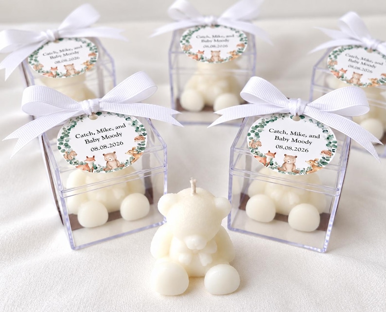 May include: Five off-white bear-shaped candles, four of which are enclosed in clear square plastic boxes topped with white ribbon bows and circular tags featuring woodland animal illustrations and the text Catch, Mike, and Baby Moody 08.08.2026.
