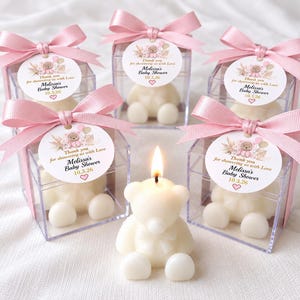 May include: Five clear square boxes containing white bear-shaped candles, each topped with a pink satin ribbon and a round tag reading Thank you for showering us with Love, Melissa's Baby Shower, 10.3.26. One lit bear candle sits in front.