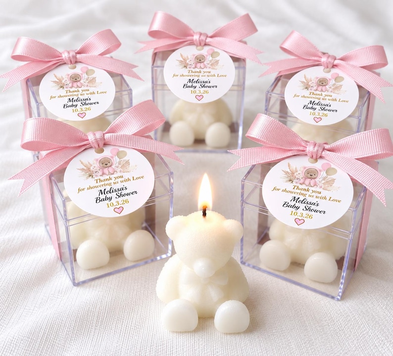 May include: Five clear square boxes containing white bear-shaped candles, each topped with a pink satin ribbon and a round tag reading Thank you for showering us with Love, Melissa's Baby Shower, 10.3.26. One lit bear candle sits in front.