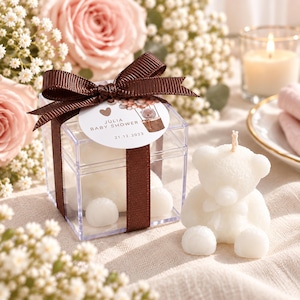 May include: A clear square plastic box containing a white bear-shaped candle, tied with a brown ribbon and a round tag reading Julia Baby Shower 21.12.2023. A matching white bear-shaped candle sits outside the box, featuring a central wick.
